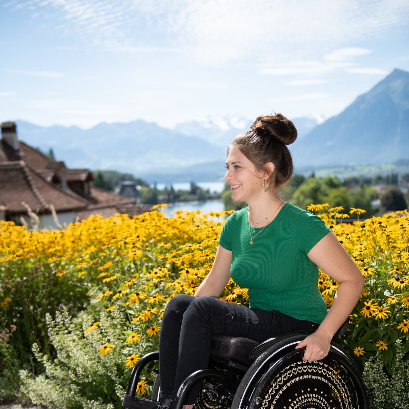 Eine junge Frau sitzt im Rollstuhl bei hellem Sonnenlicht vor einer Wiese aus gelben Blumen. Im Hintergrund sind Hausdächer, ein See und Berge zu sehen.